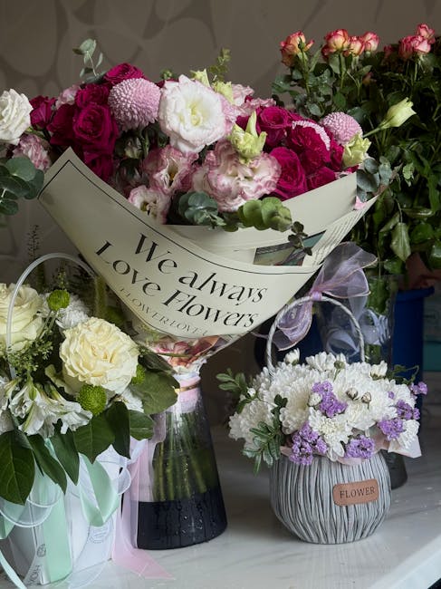 A diverse arrangement of fresh floral bouquets displayed on a surface. The central bouquet features a mix of pink and white roses, peonies, and carnations wrapped in decorative paper with the phrase '