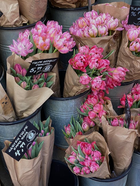 A display of fresh pink tulips arranged in brown kraft paper wraps, each bouquet placed inside galvanized metal buckets. The tulips vary in shades of pink, with some showing a delicate blush tone whil