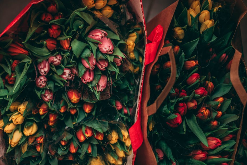 A collection of multiple bouquets of fresh tulips, predominantly in shades of pink, yellow, and red, arranged in brown paper wrap with red ribbons. The tulips have tightly closed buds with green stems