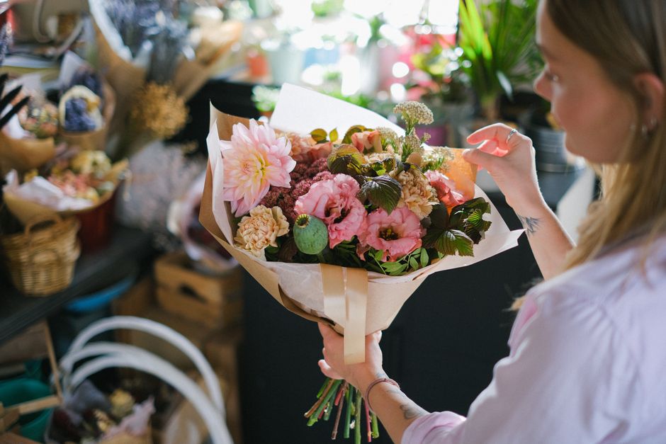 A woman with light brown hair and a tattoo on her wrist is holding a bouquet of fresh flowers wrapped in beige paper and tied with a satin ribbon, featuring pink dahlias, pale pink roses, purple lisia