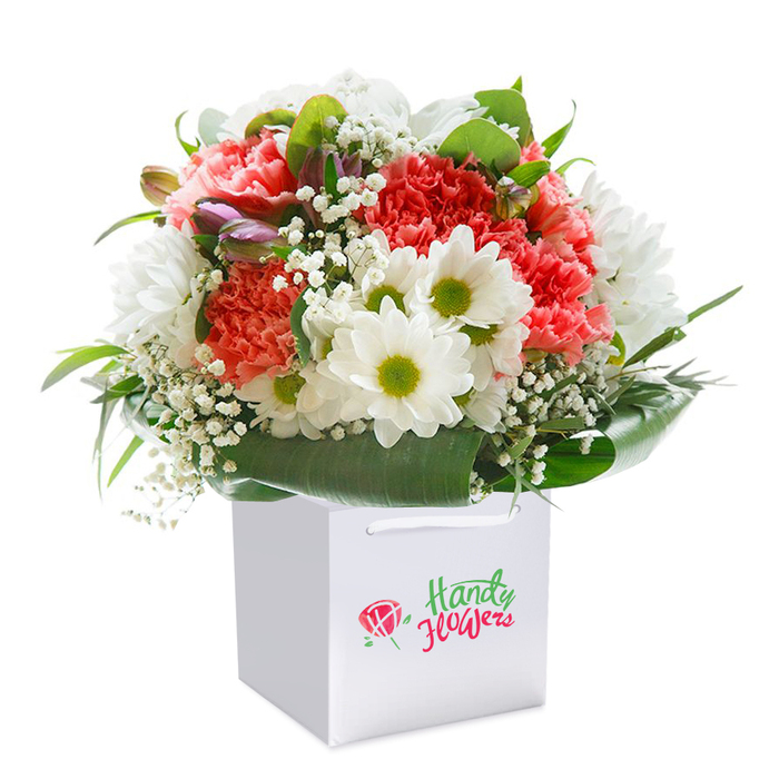 Exquisitely arranged and recalling the welcoming neighbourhoods of Chase Cross, this abundant floral bouquet stands upright in a sharp white square gift bag designed for easy carrying and presentation. Each carnation-a blend of soft coral, peach, and fiery red-bursts with layers of frilled, velvety petals, their textures creating a sense of cheerful abundance. Dotted between the carnations, clusters of bright white daisies with lively yellow-green centres capture the spirit of sunny days in local parks, while baby's breath weaves a delicate, cloud-like veil throughout. Flecks of purple from a few unopened buds add nuance and depth, anchoring the eye and echoing the diversity of local wildflowers near Chase Cross Fields. The bouquet is encircled by deep green, glossy foliage, which forms a supportive base and imparts a natural richness to the overall composition. All of this is cradled within a structured, pristine white gift bag with classic ribbon handles, its simple silhouette providing both elegance and practicality for flower delivery. Carefully styled into a rounded, harmonious arrangement, the bouquet projects freshness, quality, and ready-to-gift appeal-ideal for marking any significant day in the RM5 area, from congratulatory milestones to heartfelt remembrances.
