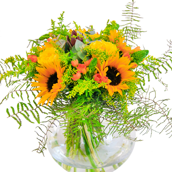 Overflowing with vivid color and energy, this Chase Cross bouquet is a true celebration of local floral artistry, housed in a round glass vase that highlights every detail. At the forefront, bold sunflowers burst open, their golden petals nearly glowing in the clear morning light-echoes of the cheerful blooms spotted along Havering Road. Nestled closely, creamy yellow garden roses spiral with refined elegance, each petal velvety soft and handsomely unfurled. Here and there, jewel-bright red hypericum berries offer a brilliant counterpoint to the yellows and greens, their gloss reminiscent of ripe fruit after a summer rain. Textural depth abounds, from the feathery sweep of ferns to the dainty spikes of filler flowers and the subtle haze of eucalyptus, lending a modern, wild-meadow appeal to the piece. The transparent vase grounds the display with its simple, clean design, letting the stems' green lines and the shimmering water within add to the bouquet's organic beauty. Bringing together freshness, artistry, and a dash of countryside whimsy, this arrangement is perfect for moments of joy-whether placed in a sunny kitchen or delivered as a heartfelt gift across Chase Cross and its welcoming neighbourhoods.
