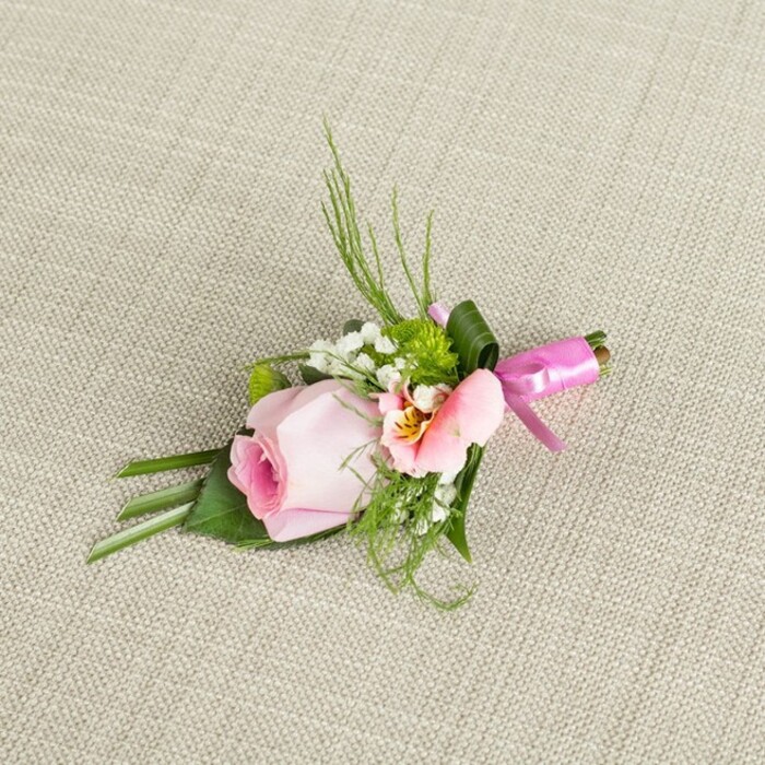 Gracefully positioned on a lightly textured woven linen-so typical of elegant Chase Cross banquets-this boutonniere is a testament to local floral talent. Its focal point: a tightly spiraled, soft pink rose with a gentle ombré from lush blush at the core to paler outer petals, conjuring memories of wandering through Chase Cross's green spaces in May. A dainty blush-peach alstroemeria flower, with open petals marked by delicate yellow and subtle veins, complements the central rose and introduces a nuanced splash of colour. Plumes of white baby's breath drift airily around the central cluster, their small blooms reminiscent of the morning mist over Bedfords Park. Green miniature cushion mums dot the design, playfully textured amid the wispy backdrop. The arrangement is interwoven with leafy details-long, dark green blades rising upwards, wide flat leaves cradling the flowers at the base, and a single broad leaf embracing the clustered stems. The boutonniere is united with a vibrant fuchsia pink satin ribbon, tied as a crisp bow that gleams with celebratory flair. Whether destined for a wedding, prom, or a cherished family gathering at a Chase Cross venue, this floral accessory blends romance, freshness, local colour, and artisanal skill into a single beautiful piece.